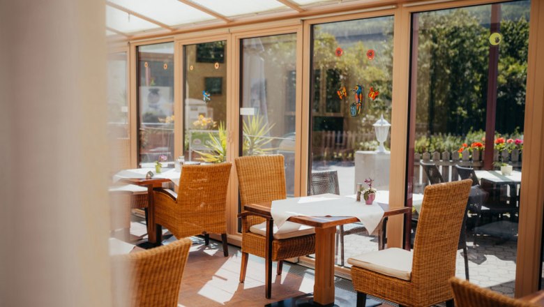 A bright conservatory with wicker chairs and laid tables, surrounded by large windows with a view of the greenery.