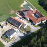 Aerial view of a farm with several buildings and solar panels on a roof, surrounded by fields and forests.