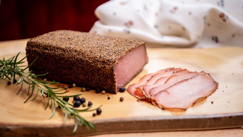 A piece of cured meat with pepper crust and sliced on a wooden board, decorated with rosemary and juniper berries.