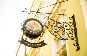 Metal sign 'Café Konditorei Hagmann' on a yellow house wall.
