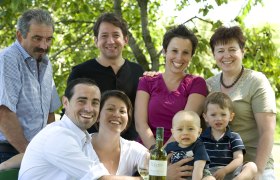 A smiling family poses outdoors with a bottle of wine on a table.