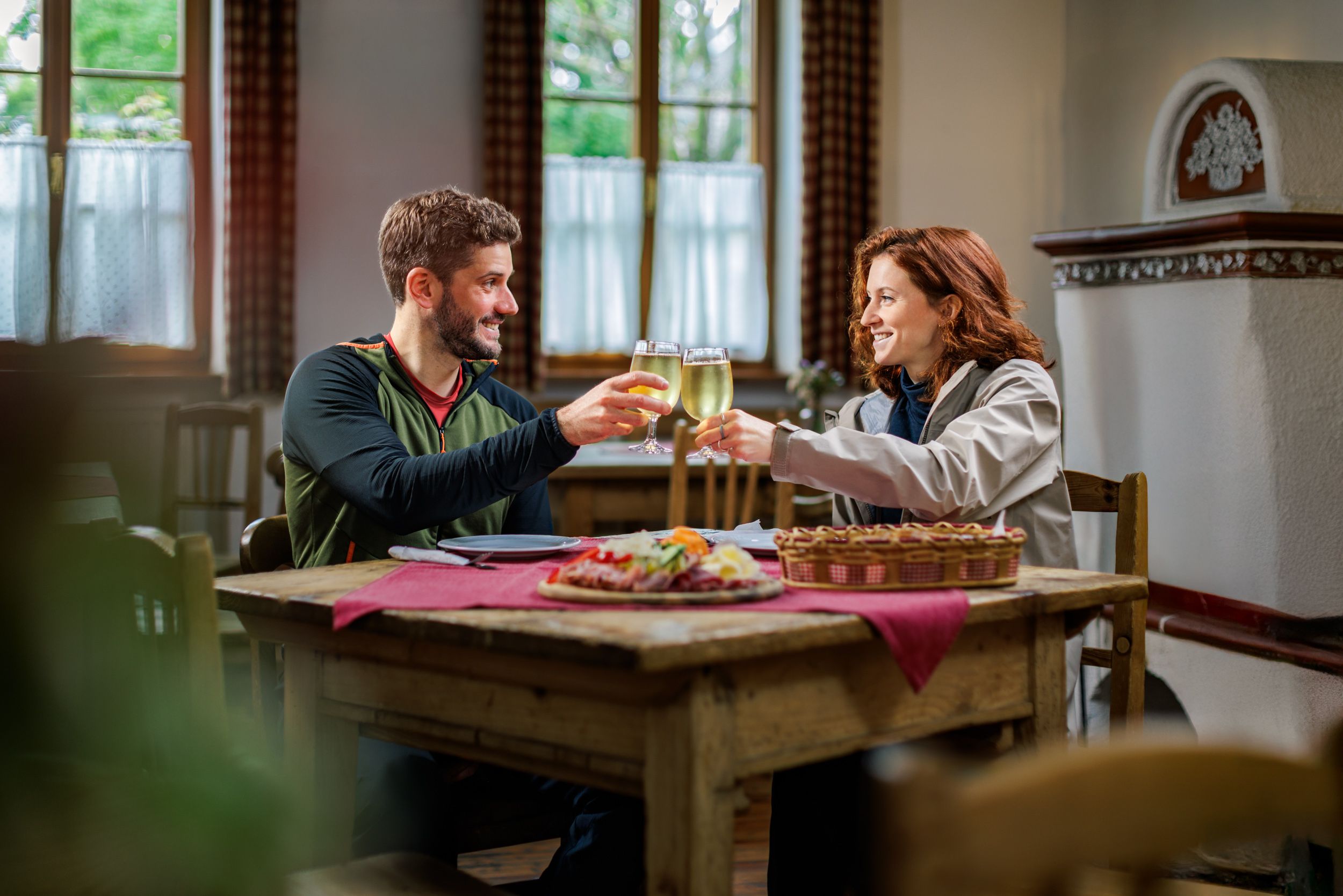 A couple sits in a parlor on a rustic wooden table in front of a Brettljause and toasts with apple cider