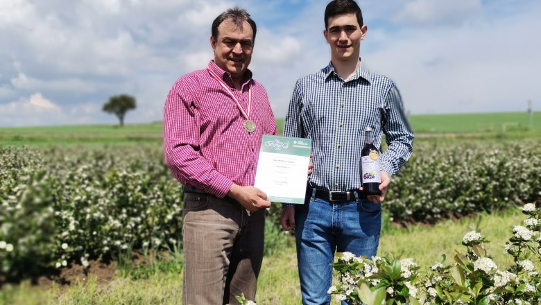 Two men stand in a flowering field, one holding a certificate and a medal, the other a bottle.