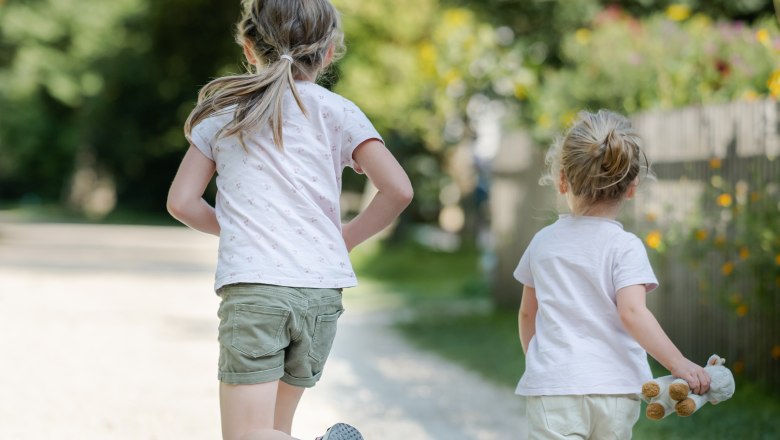 Two children are running on a gravel path in a garden, surrounded by trees and flowers.
