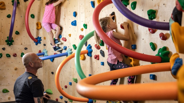 Children climb on a colorful climbing wall with foam rings while an adult supervises.