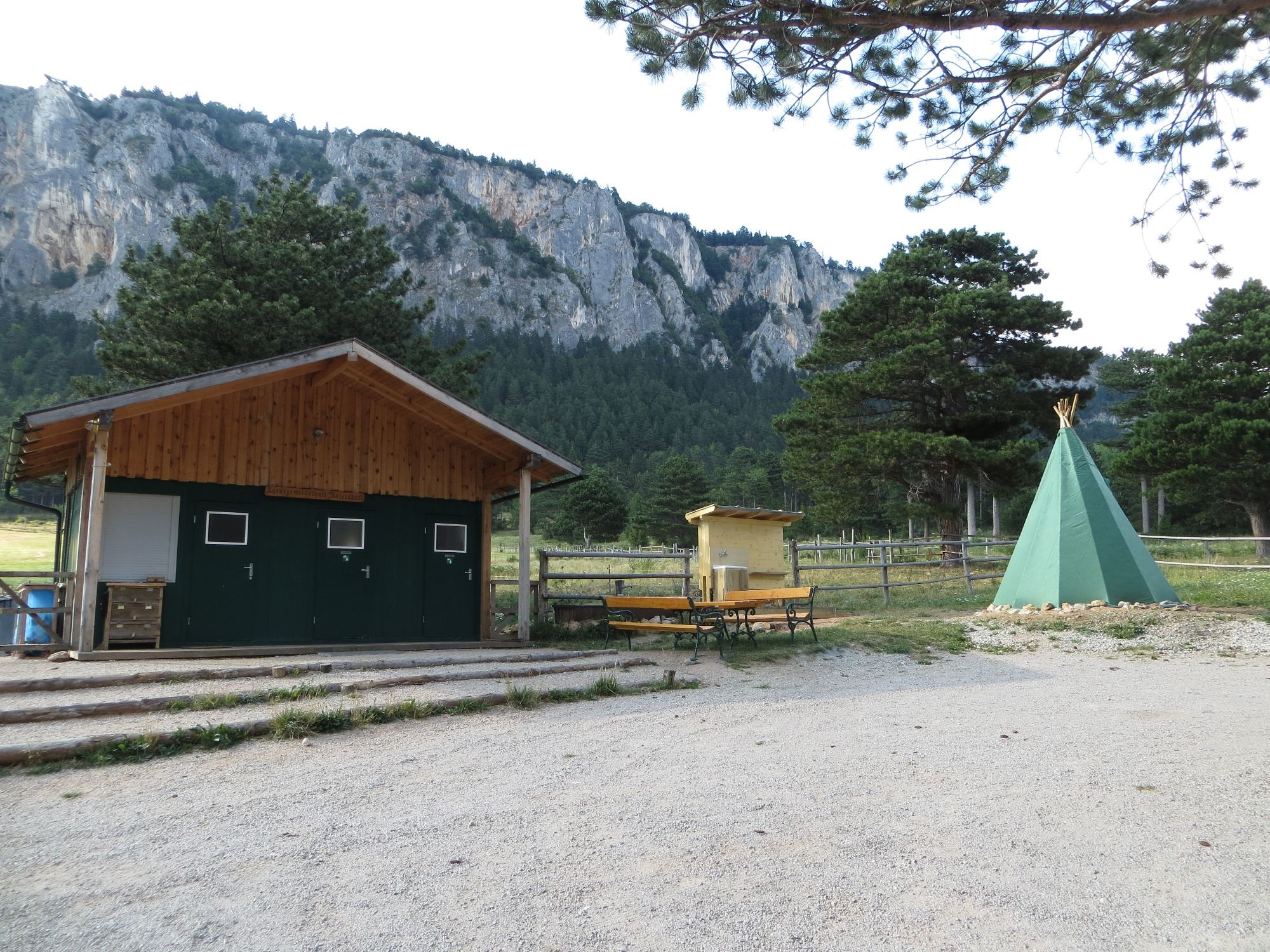 A campsite with a green tepee, a wooden hut and benches in front of a rock face and trees.