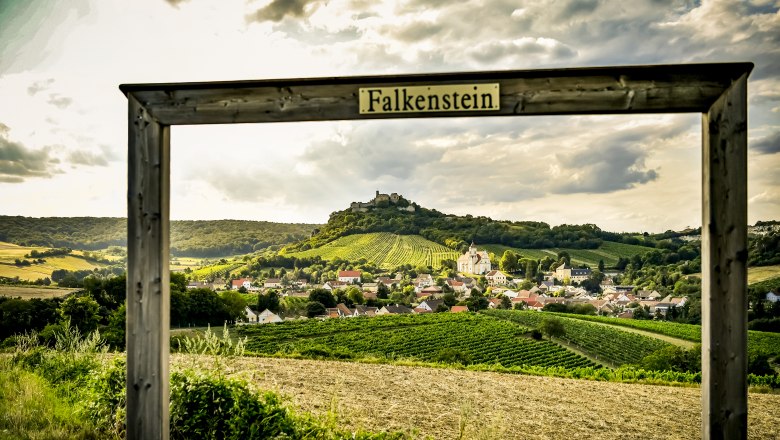 View of the village of Falkenstein with vineyards and a ruined castle in the background, framed by a wooden frame with the inscription 'Falkenstein'.