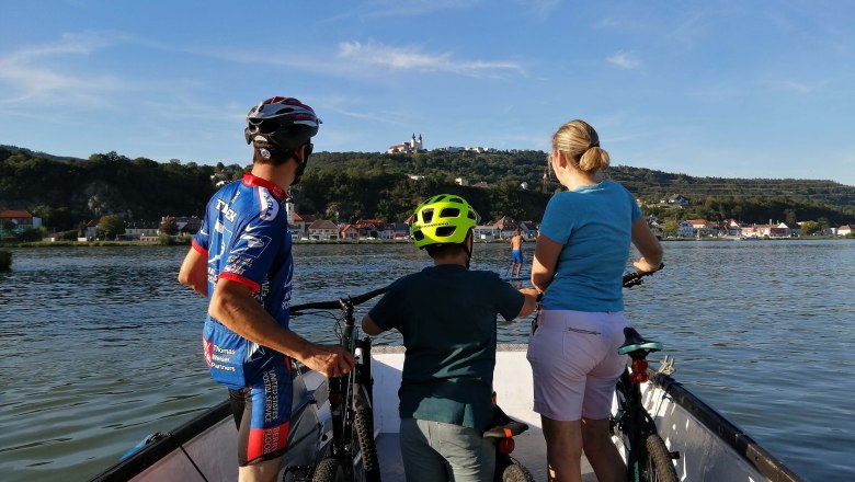 Three people with bicycles on a boat crossing a river. A town and a hill with a building can be seen in the background.