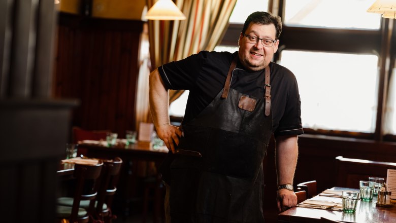 Man in a black apron stands smiling in a cozy restaurant.