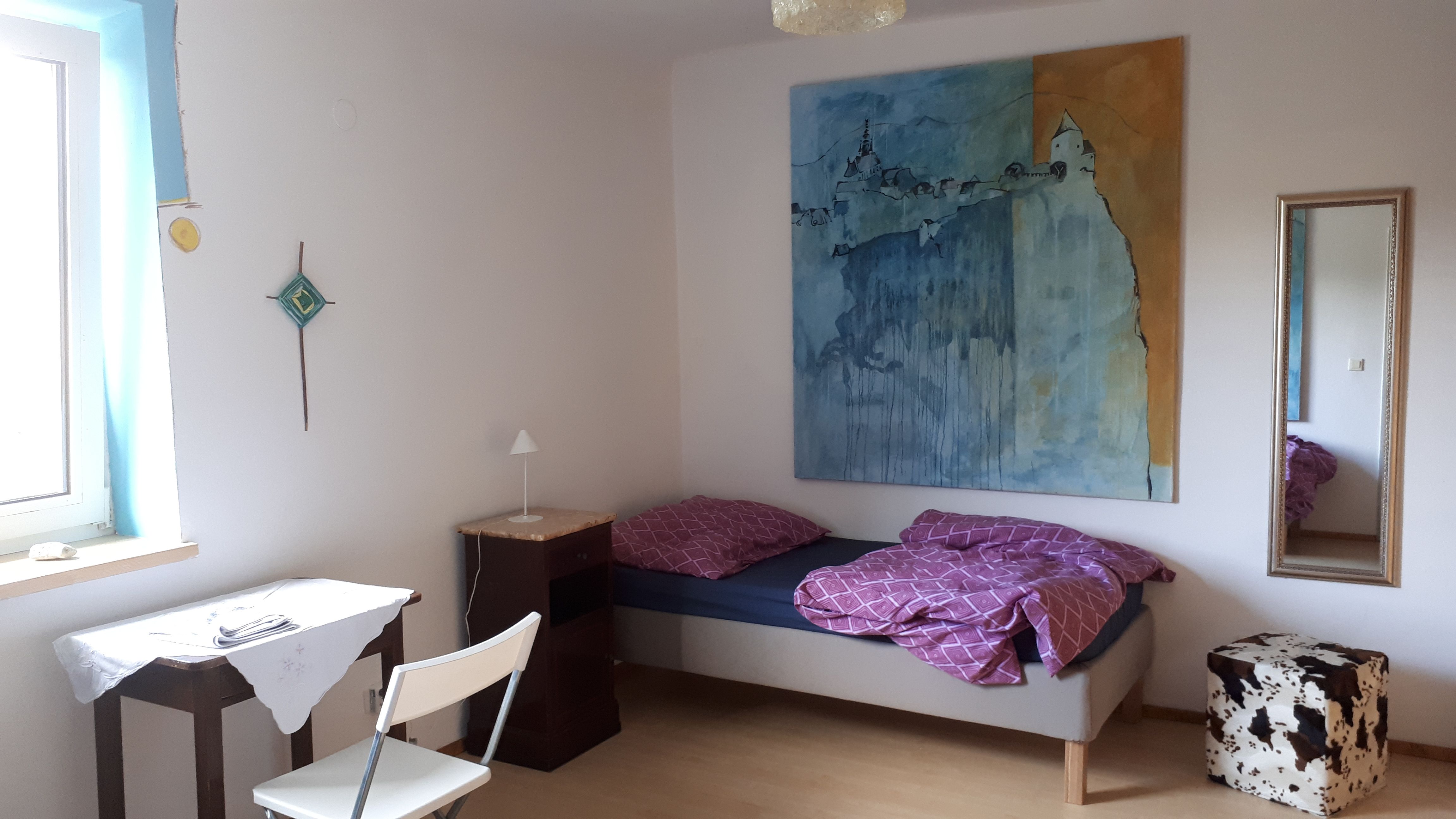 A simply furnished room with a bed, table, chair and large painting on the wall.
