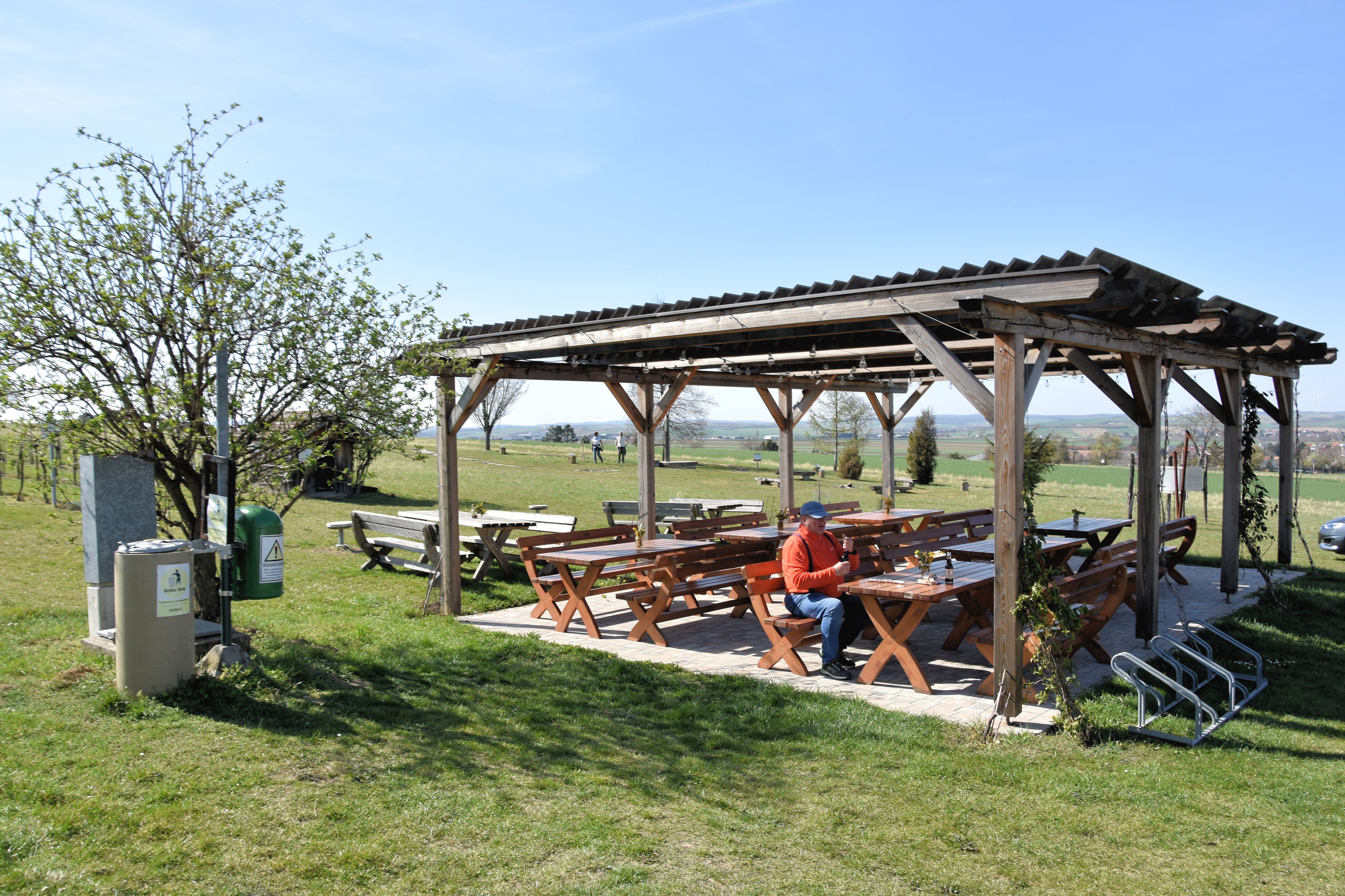A covered rest area with wooden benches and tables on a meadow, a person is sitting there. Fields and trees can be seen in the background.