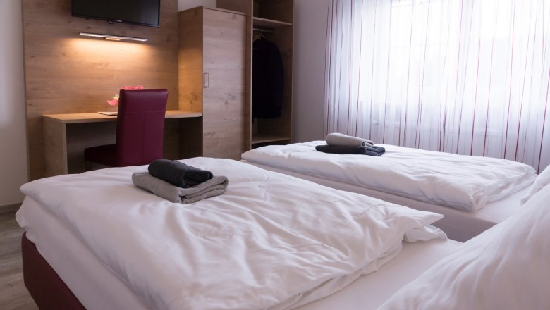 Twin room with two beds, desk, chair and closet. Towels on the beds, window with curtains in the background.