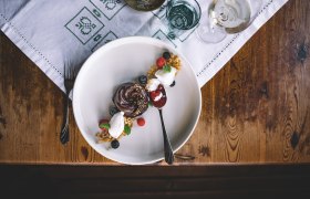 A dessert plate with a nougat tartlet, elderberry ice cream, berries and a spoon on a wooden table.