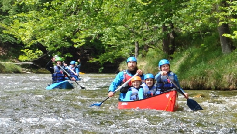 People in canoes paddling on a river in a green forest landscape.