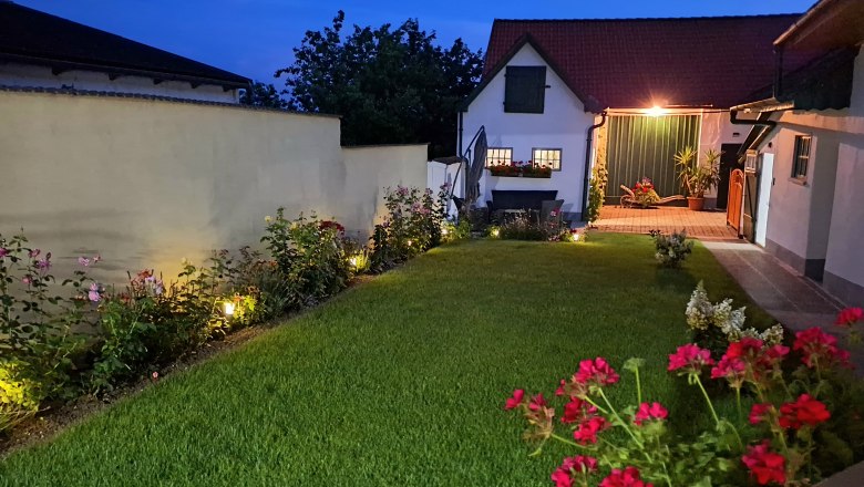 An illuminated courtyard at night with a manicured lawn, flower beds and a small building in the background.