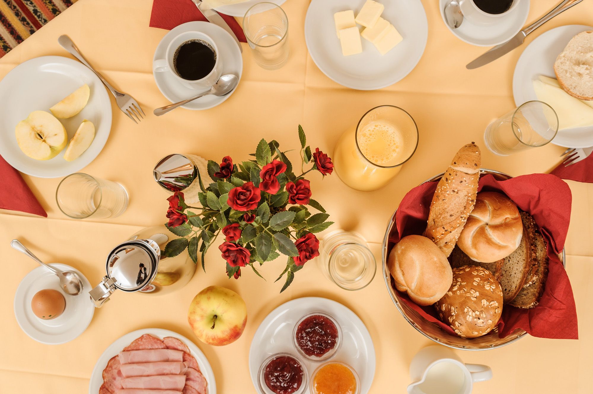 A laid breakfast table with bread rolls, cold cuts, jam, coffee, orange juice and a bouquet of flowers in the middle.