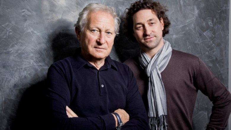 Two men stand in front of a gray wall, one older with folded arms, the other younger with a scarf.