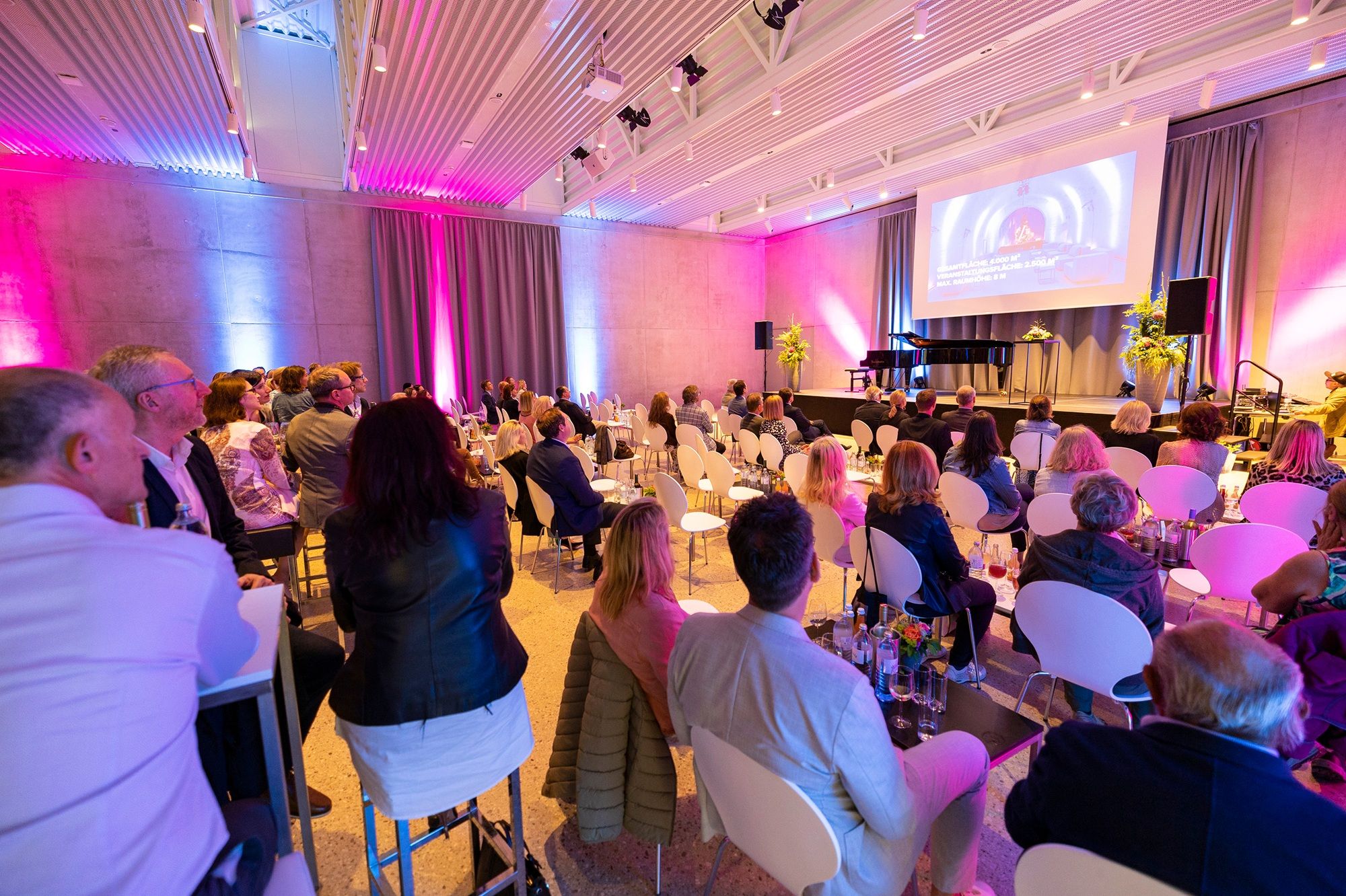People sit in a modern event room with a stage and screen.