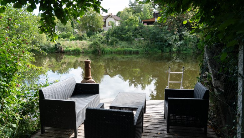 A wooden footbridge with black garden furniture on the riverbank, surrounded by green vegetation and a house in the background.