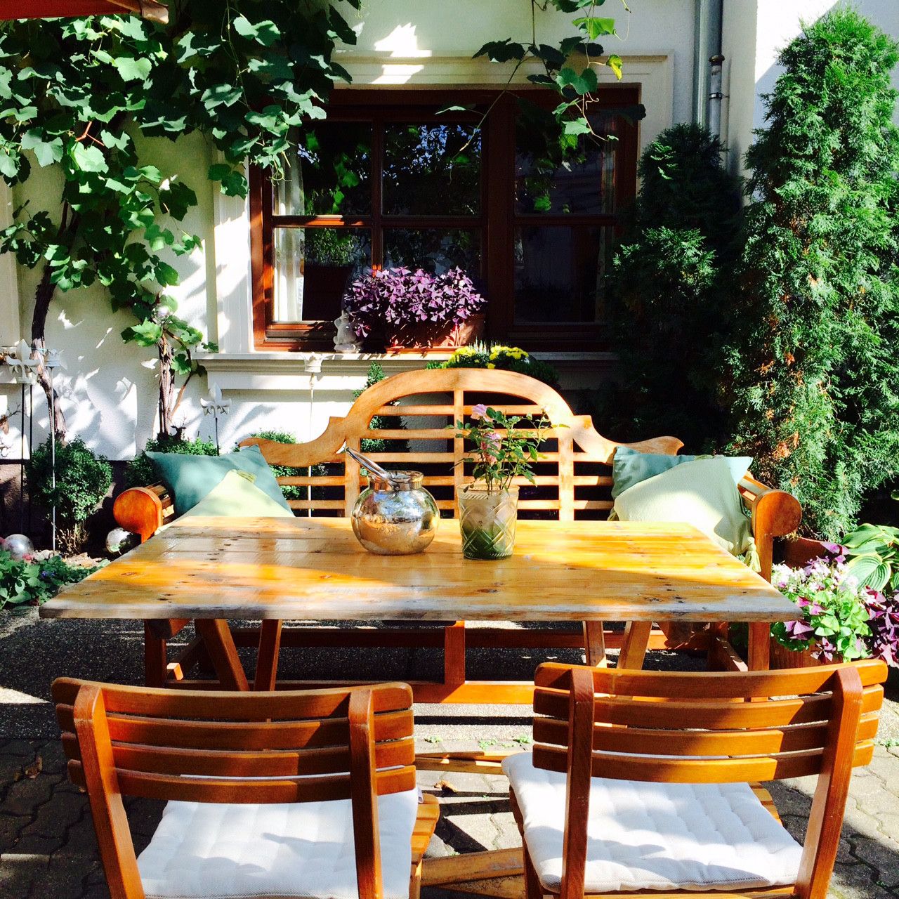 Cozy garden area with wooden table and chairs, surrounded by plants and vines.