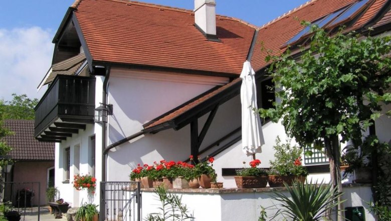 A white house with a red tiled roof, balcony and garden with plants and flowers.