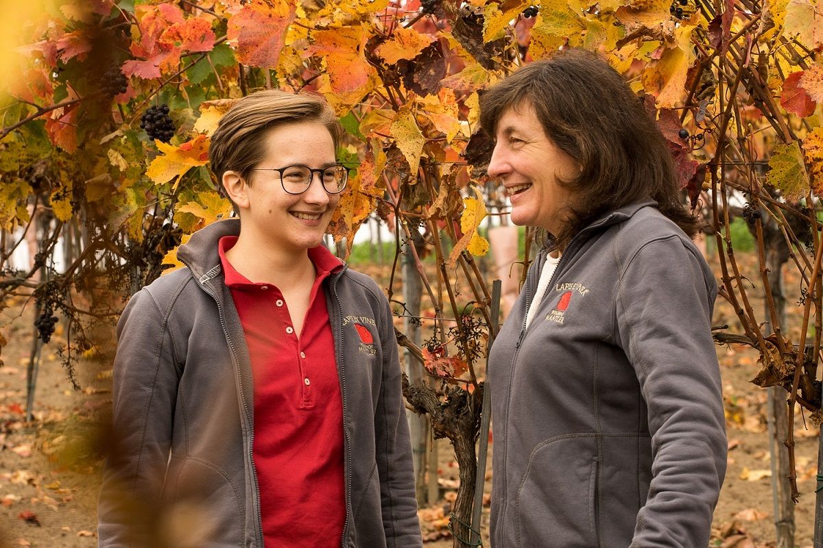 Two women stand smiling in an autumnal vineyard with colorful leaves.