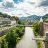Panorama of a river landscape with a castle and modern architecture, surrounded by green hills.