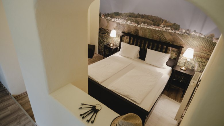 A cozy bedroom with a double bed, two bedside lamps and a wallpaper with a wine landscape.