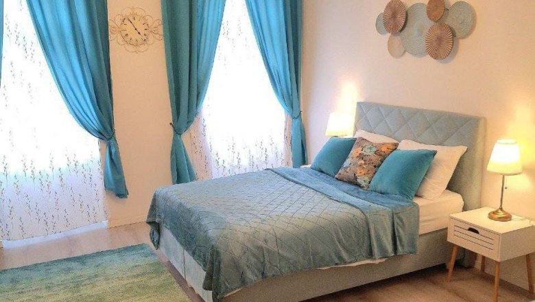 A cozy bedroom with blue curtains, a double bed and decorative cushions. Two bedside lamps illuminate the room.