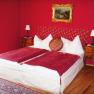 A bedroom with red walls, a double bed with red blankets and a painting on the wall.