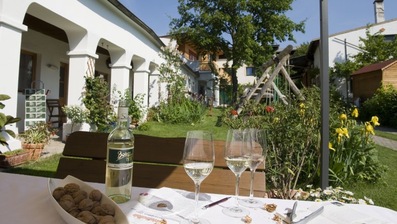 An idyllic courtyard with a table, wine bottle, glasses and nuts in the foreground. In the background, a garden with flowers and a tree.