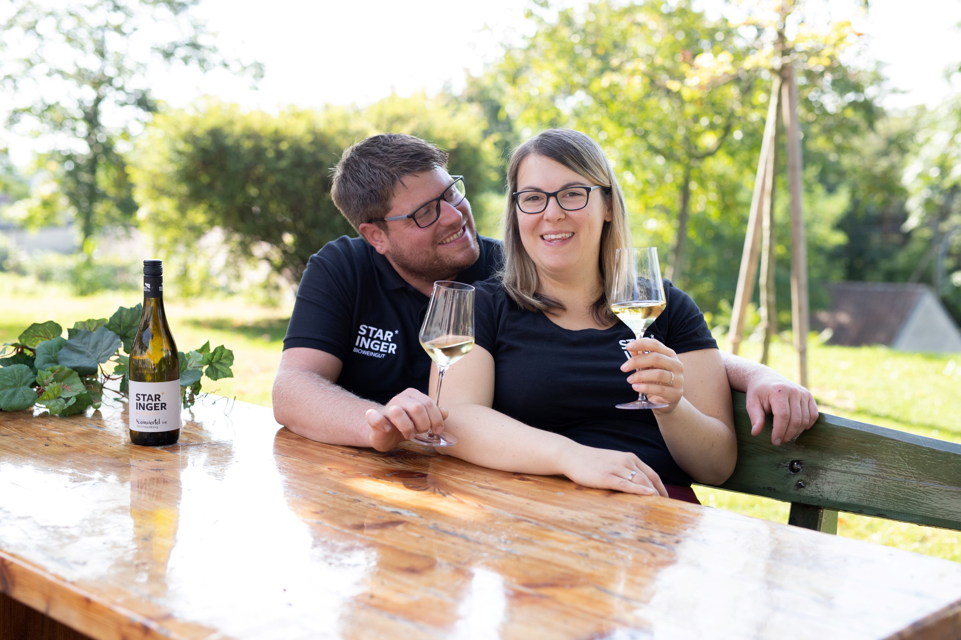A couple is sitting at a wooden table outside, holding glasses of wine. A bottle of wine with the label 'Staringer' is on the table.