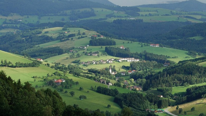 Panoramic view of a green, hilly landscape with scattered houses and forests.