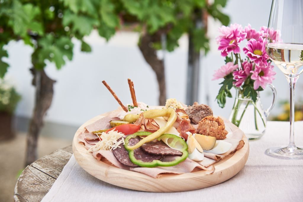 A platter snack with various types of sausage and cheese, bread and vegetables on a wooden plate, with a glass of white wine and flowers next to it.