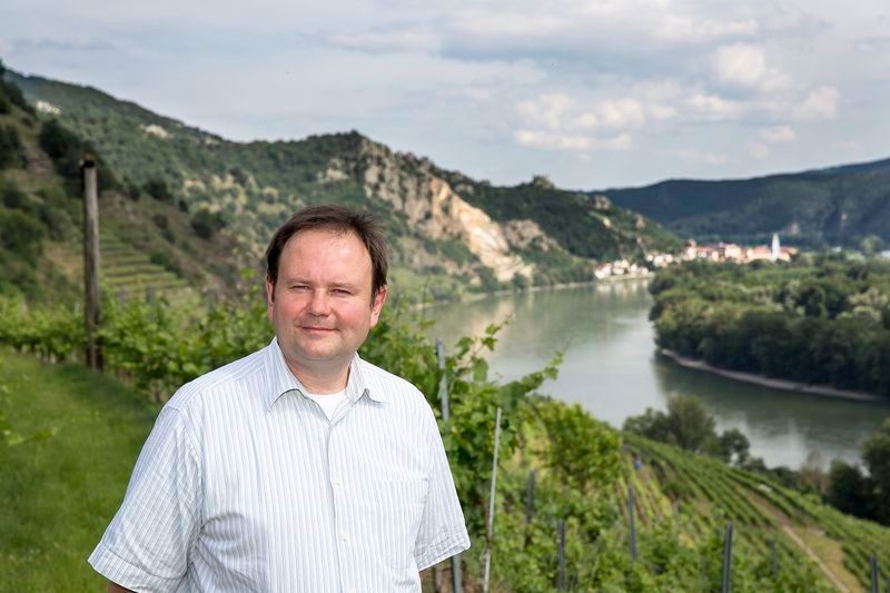 Man standing in a vineyard with river and hills in the background.