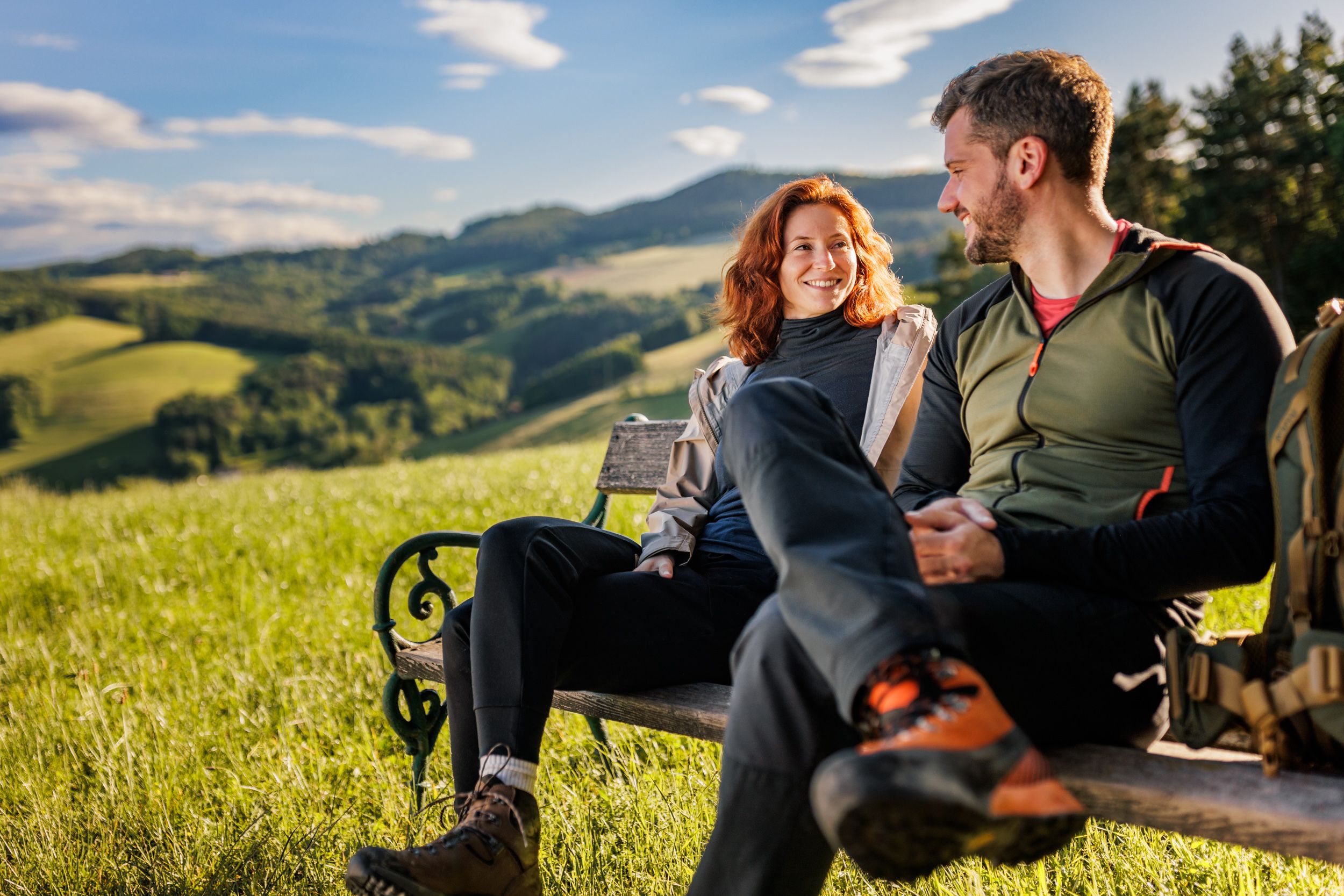A woman and a man in hiking gear are sitting on a wooden bench. Meadow, green hills and forest in the background.