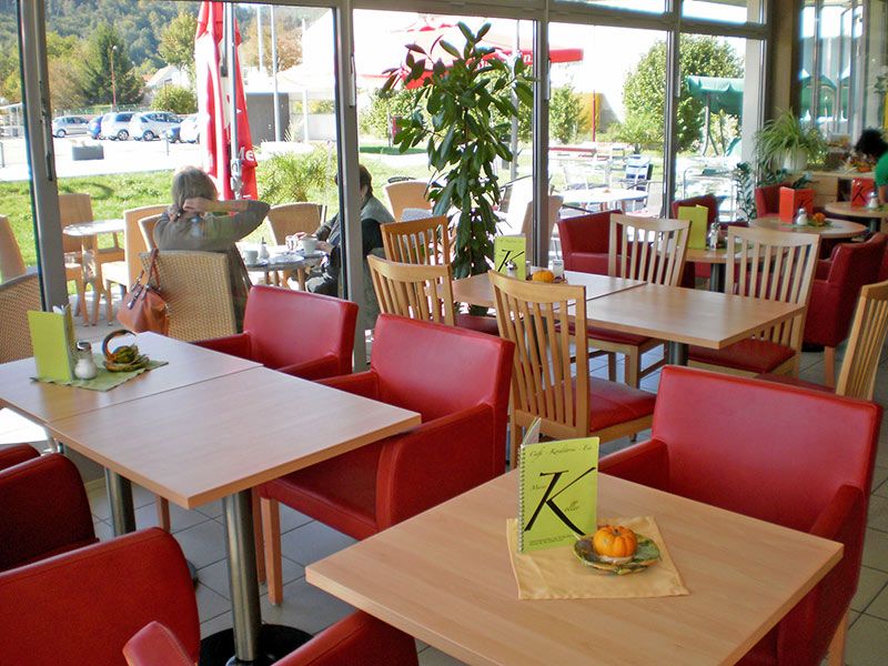 Interior view of a café with red armchairs and wooden tables, view of terrace with parasols.