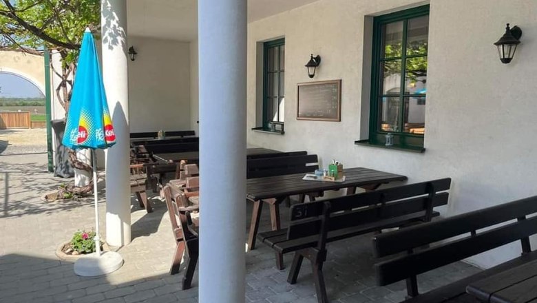 An empty guest garden with wooden benches and tables, a parasol and a white wall with windows.