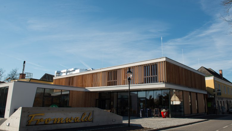 The Hubertushof in Bad Fischau-Brunn, © Fotografin: Eva Heindl Modern building with wooden façade and glass front, in front of it a sign with the inscription 'Fromwald'.