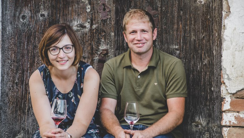 A man and a woman are sitting in front of a wooden wall, holding wine glasses in their hands.