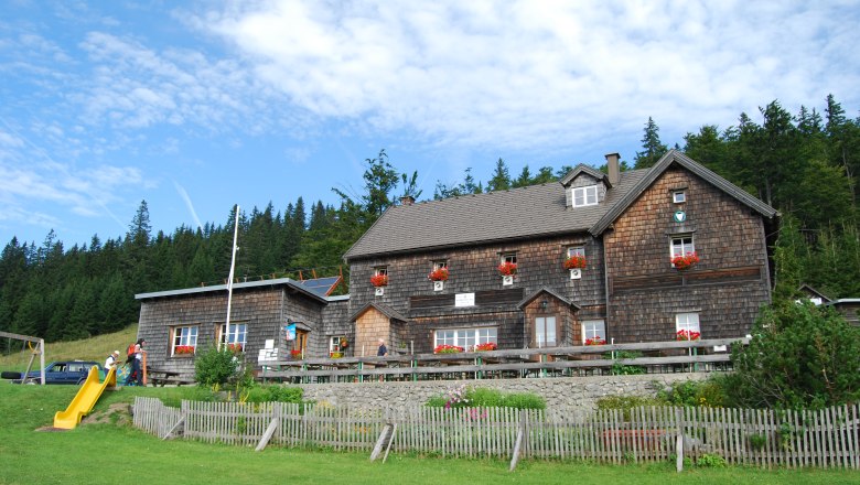 A traditional wooden house with red flower boxes in front of a forest and blue sky.