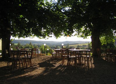 A shady garden with wooden tables and chairs under large trees, with a view of a hilly landscape in the background.