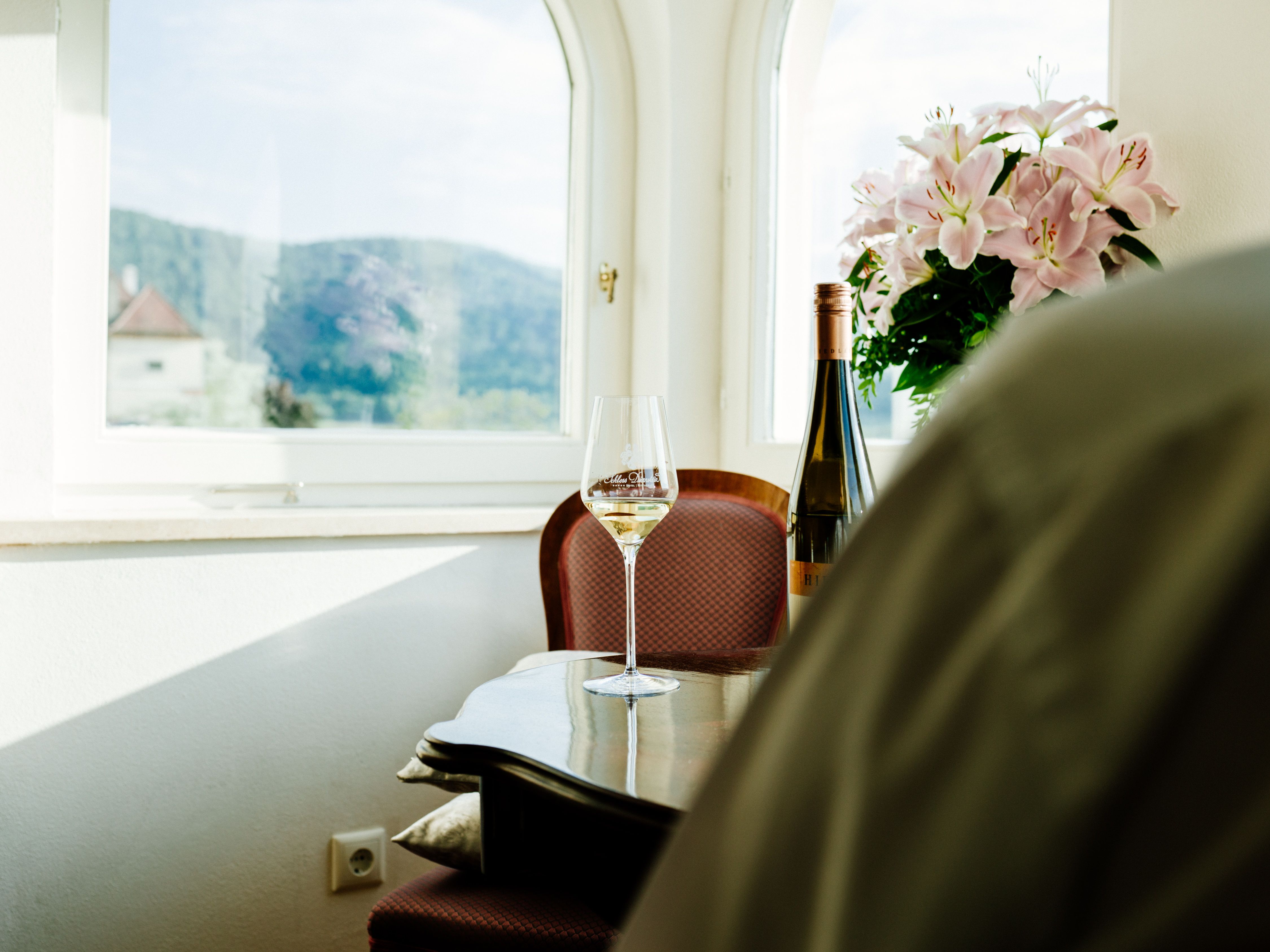 A wine glass and a bottle of wine on a table in front of a window with a view of hills and flowers.