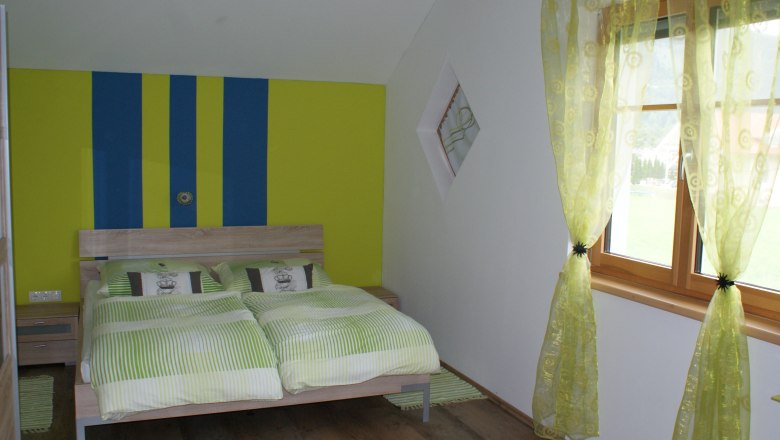 Bedroom with double bed, green and yellow striped wall, green curtains, wooden floor.