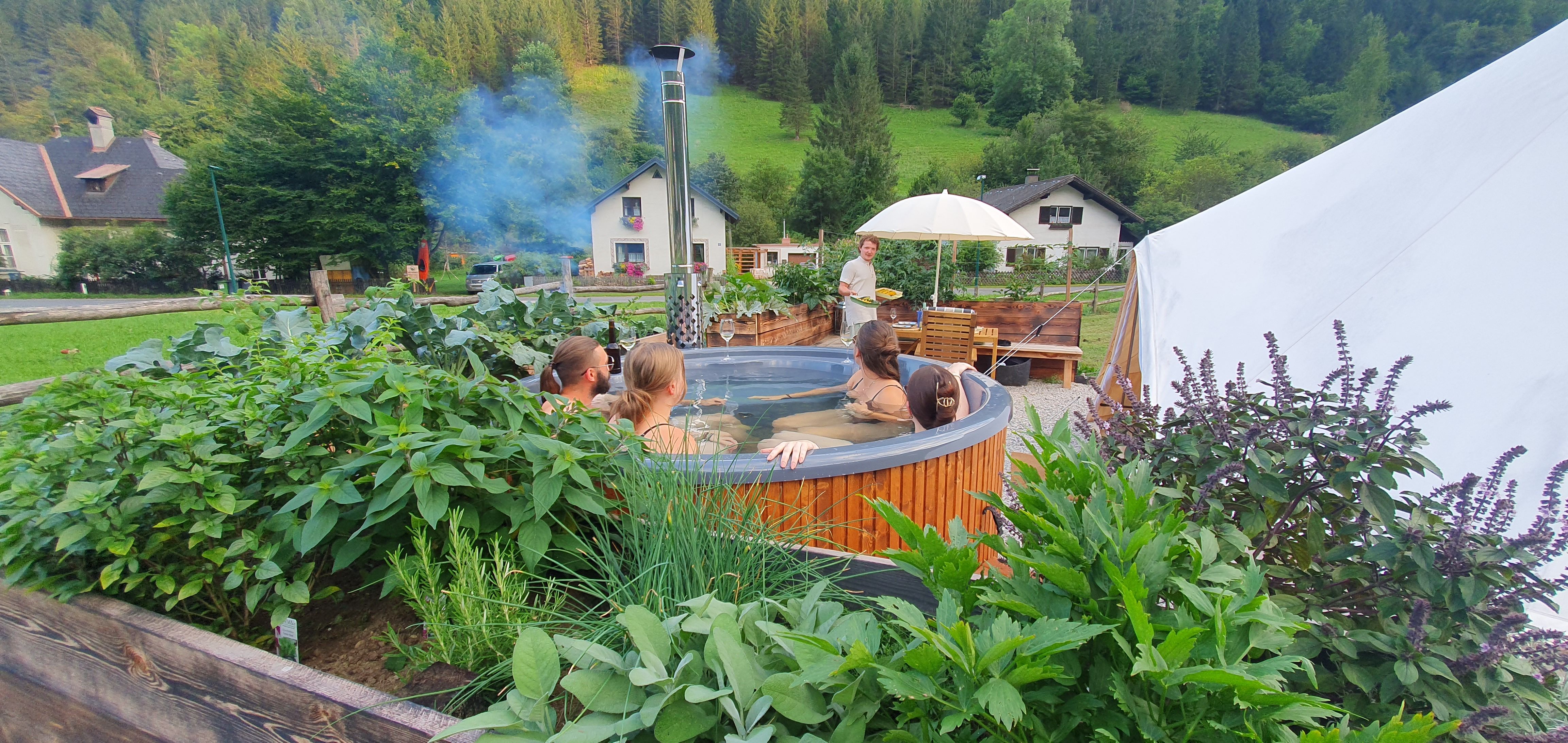 People in a round wooden whirlpool surrounded by plants, with a tent and houses in the background.