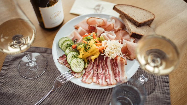 A plate of cold cuts, vegetables and bread, surrounded by wine glasses and a bottle of wine on a table.
