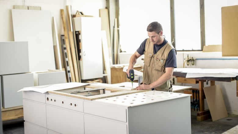 A craftsman is working on a piece of furniture in a workshop with a cordless screwdriver.