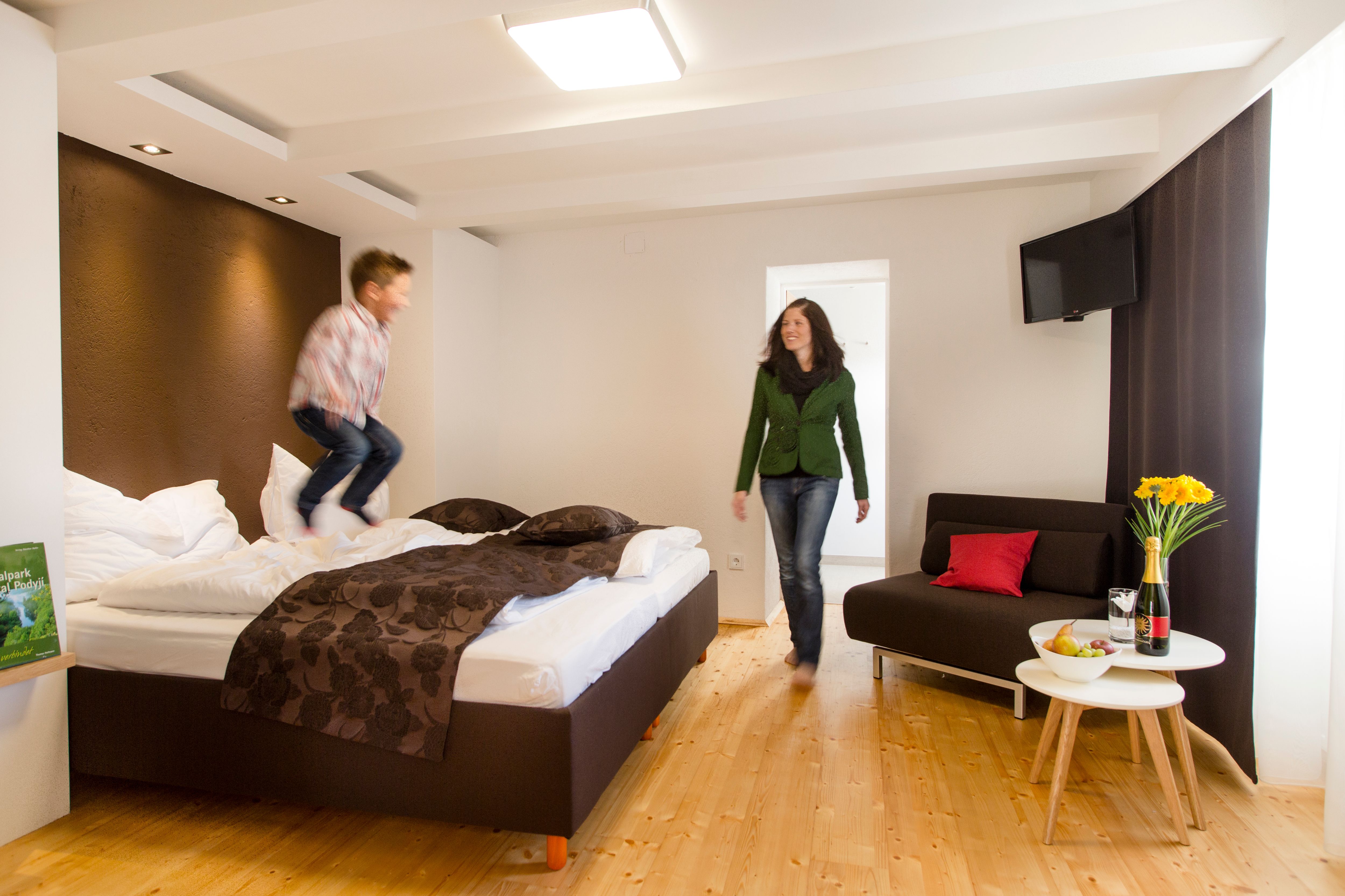 A child jumps on a bed in a modern hotel room while a woman looks on smiling. A table with flowers and wine stands next to it.