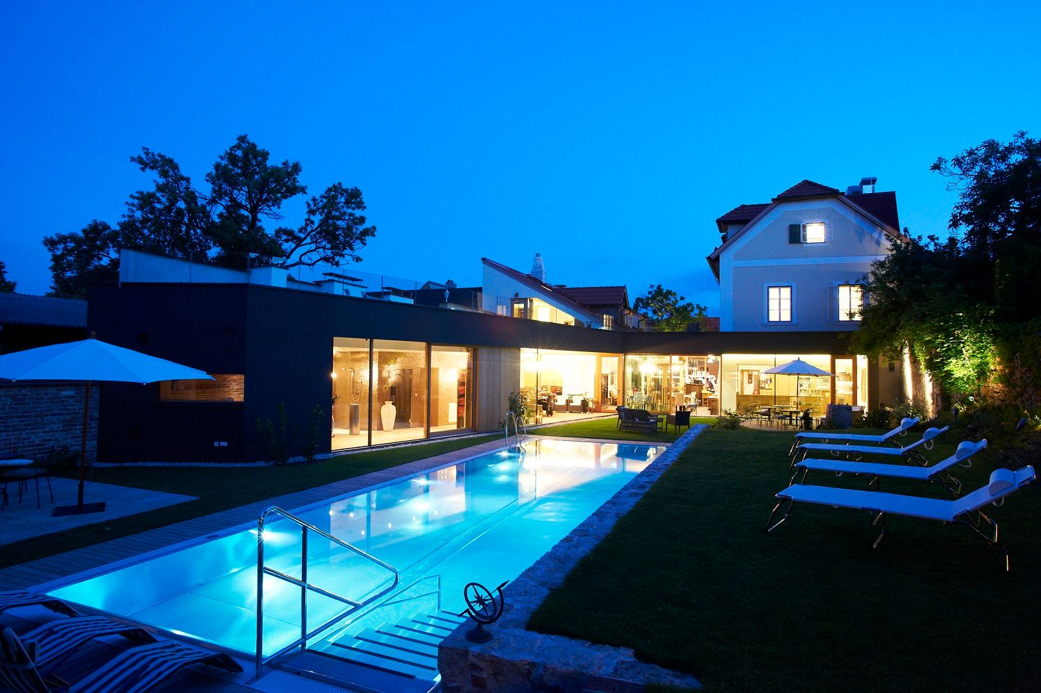 Evening view of a modern hotel with illuminated pool and sun loungers in the garden.