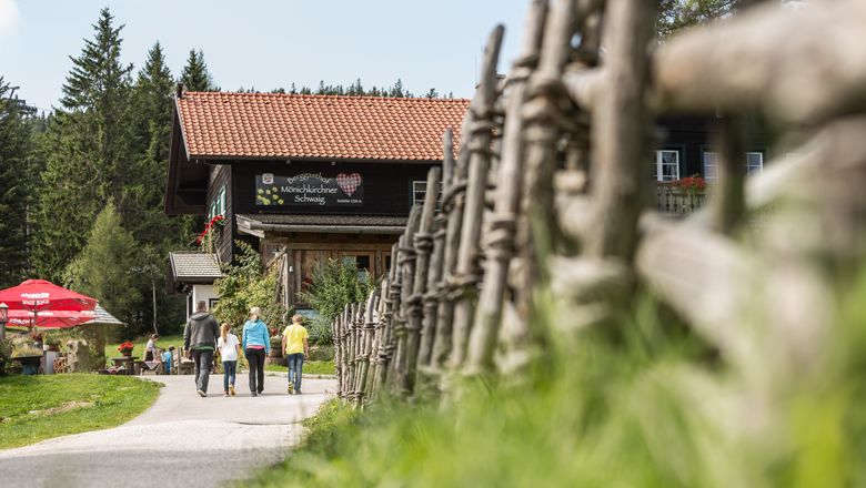 Three people are walking towards a mountain inn, surrounded by a wooden fence and trees.